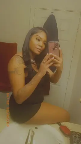 Young woman in a black dress taking a mirror selfie in a bathroom with a moody, warm aesthetic.