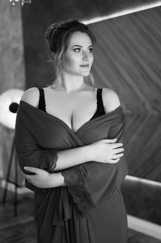 Bustybeauty18 poses against a white background, wearing a black long-sleeved top that shows deep cleavage, with her blonde hair styled down.