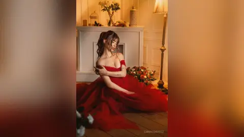 Elegant woman in a flowing red gown sitting in a warm, moody indoor setting with floral decor.
