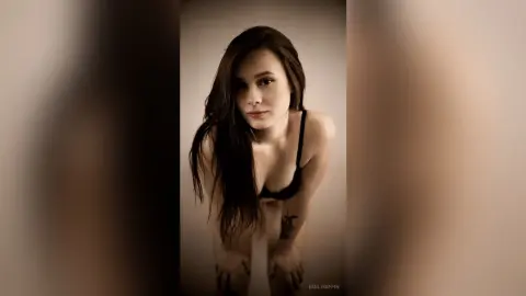 EmilyGiffin poses leaning forward in a black bra, looking directly at the camera with her long dark hair falling over her shoulder.