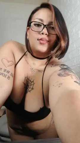 Smiling woman with glasses and tattoos wearing a black bikini in a bright indoor setting.