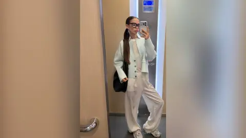 CarlynKernell poses in a mirror wearing a light blue button-up cardigan, a white top, and white trousers with her long brown hair pulled back.