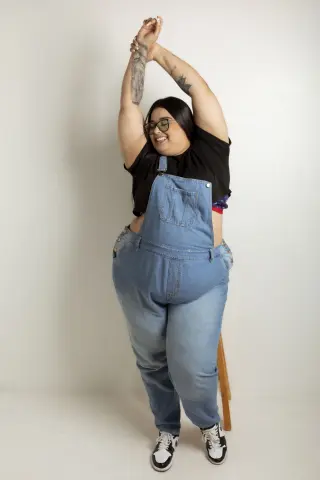 Smiling plus-size woman in denim overalls and black t-shirt posing happily against a white studio background.