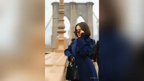 Stylish woman in a navy blue coat and sunglasses standing by the Brooklyn Bridge in a moody urban setting.