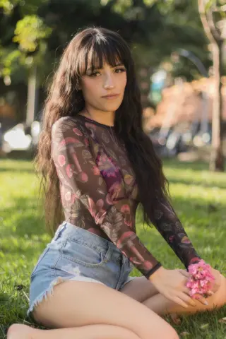 Young woman with long wavy hair wearing a floral sheer long-sleeve top and denim shorts sitting on green grass.