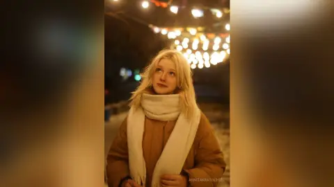 Blonde woman in a warm winter coat and white scarf looking up under glowing city lights at night.