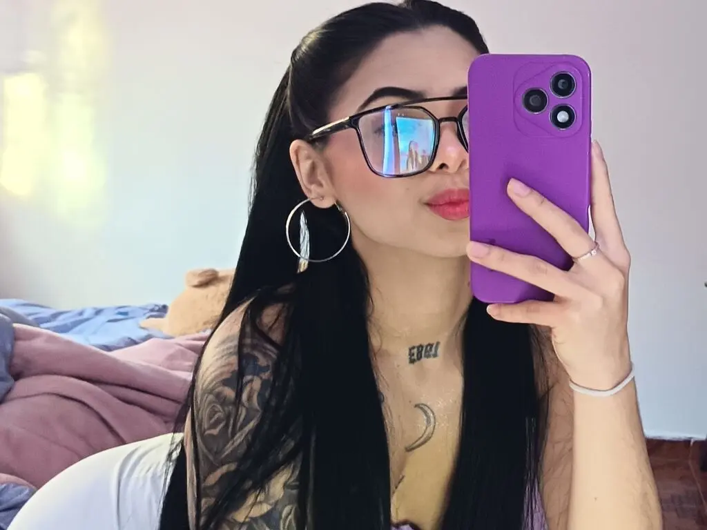 Young woman with long dark hair and glasses taking a casual mirror selfie in a cozy indoor setting.