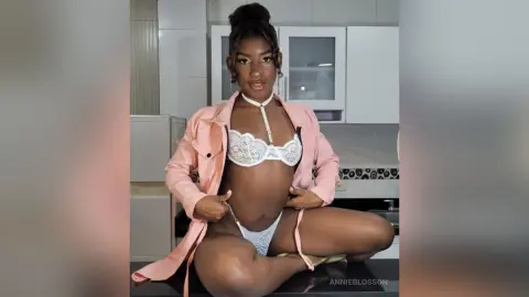 Young Black woman in white lace lingerie and a pink robe sitting in a modern kitchen with a confident mood.
