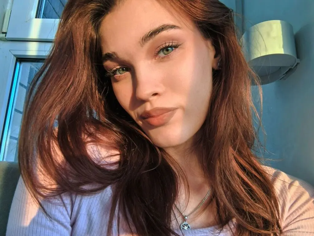 Close-up portrait of a young woman with long reddish-brown hair and green eyes in warm sunlight.