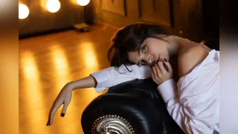 Dreamy portrait of a woman in a white shirt leaning on a black leather sofa in warm, moody lighting.