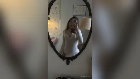 Young woman in a white long-sleeve bodysuit taking a mirror selfie in a cozy, dimly lit bedroom.