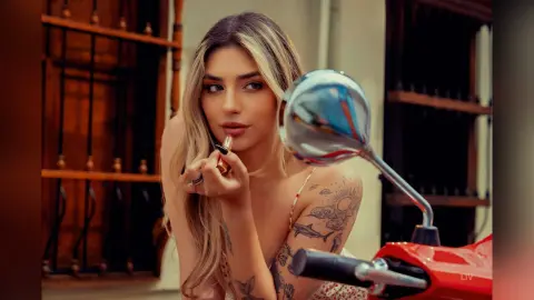 Blonde woman in a floral strap top applying lipstick while sitting on a red scooter in a warm outdoor setting.