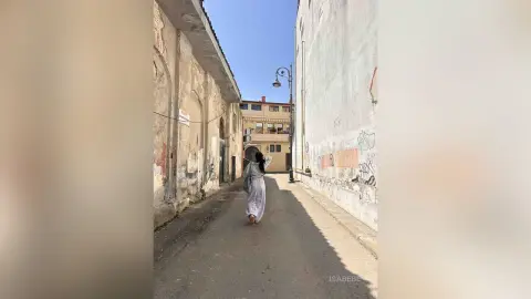 Woman in a flowing white dress walking down a narrow, sunlit European alleyway with rustic stone buildings.