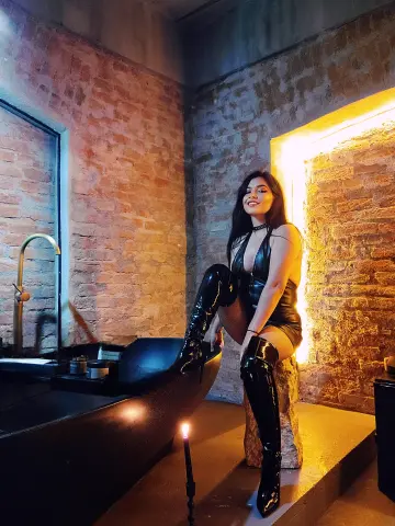 Smiling woman in a black leather outfit and thigh-high boots posing in a moody, rustic brick room with warm lighting.