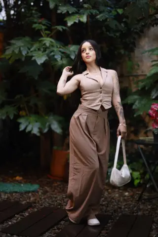 Young woman in beige linen vest and wide-leg trousers posing in a lush green garden setting.
