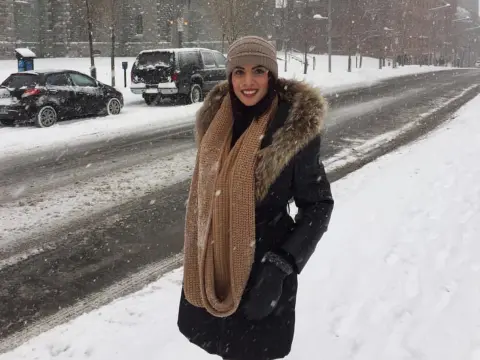 Smiling woman in a black winter coat and tan scarf standing on a snowy city street during a snowfall.