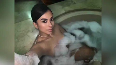VictoriaCapri relaxing in a bubble bath, showing her bare chest and shoulders with her dark hair pulled back in a sleek bun.