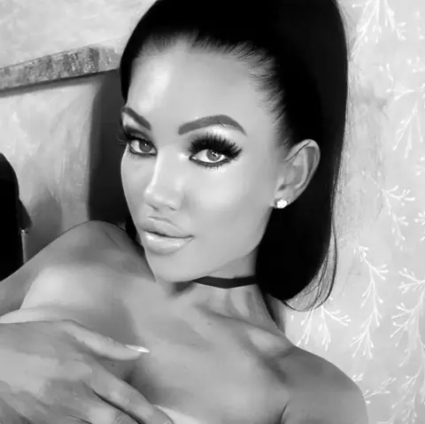 BritishTastyCherryxo poses with long dark hair and heavy lashes, looking at the camera while her hand rests on her chest.
