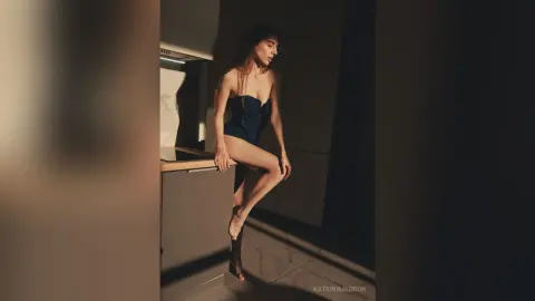 Moody portrait of a woman in a dark navy bodysuit sitting in a sunlit modern kitchen corner.