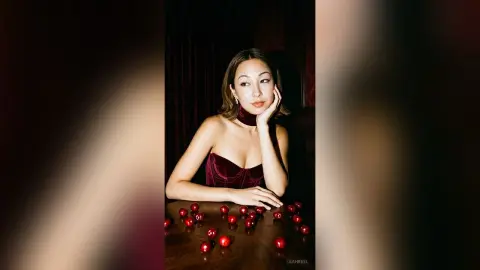 Elegant woman in a red velvet top posing thoughtfully at a table with scattered red cherries in moody lighting.