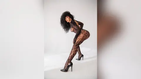 Curvy woman in a black lace bodysuit and high heels posing against a white studio background.