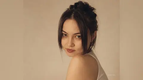 SeeBugl looks over her shoulder with dark hair pulled into a bun, wearing a white tank top against a plain background.