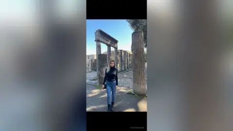 Blonde woman in a black turtleneck and blue jeans standing by ancient stone ruins under bright sunlight.