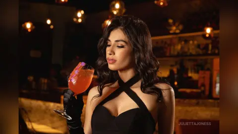 Elegant woman in a black cocktail dress and gloves sipping a drink at a moody, dimly lit upscale bar.