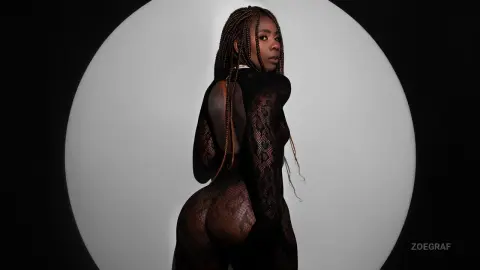 Elegant woman with long braids in a black sheer lace bodysuit posing against a minimalist circular backdrop.