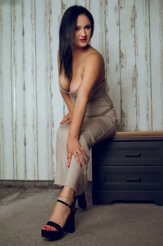 Elegant woman in a beige bodycon dress sitting on a dark dresser against a rustic white wood wall.