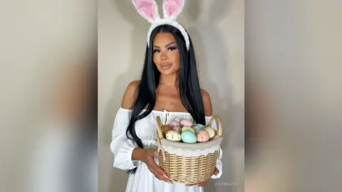 Smiling woman in white off-the-shoulder dress wearing bunny ears and holding a basket of colorful Easter eggs.
