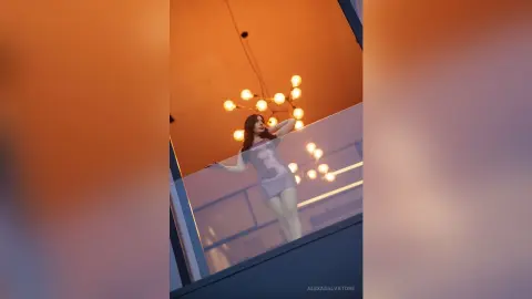 Elegant woman in a chic cocktail dress leaning on a balcony railing under warm glowing lights.