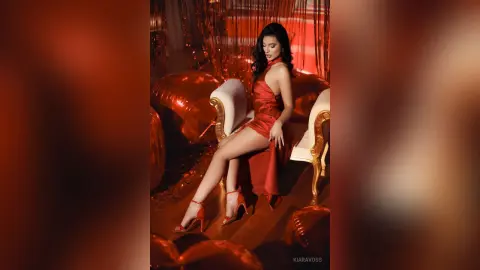 Elegant woman in a red satin dress and heels sitting on a white sofa in a glamorous red-lit room.