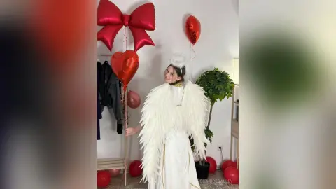 Cheerful woman in white angel costume with large wings holding red heart balloons in a festive room.
