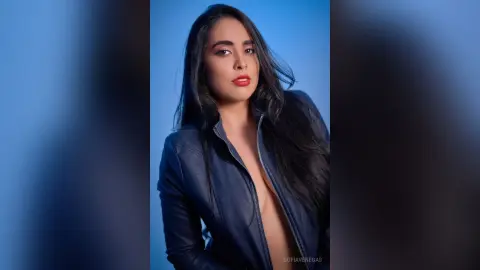 Beautiful woman with long dark hair wearing a navy blue leather jacket against a blue studio background.