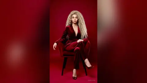 Elegant woman in a velvet burgundy jumpsuit sitting on a chair against a deep red studio background.