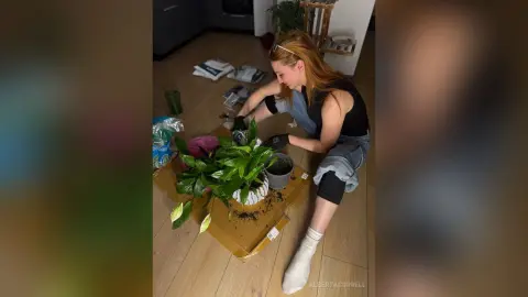 AlbertaCorrell with long red hair sits on the floor in a black tank top and denim shorts, repotting a green plant.