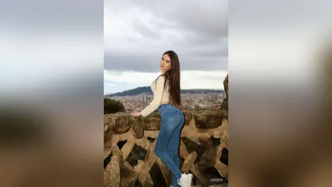 Young woman in white long-sleeve top and blue jeans posing on a stone balcony overlooking a city skyline.