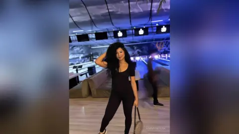 Stylish woman in a black bodysuit posing in a modern, dimly lit bowling alley lounge.