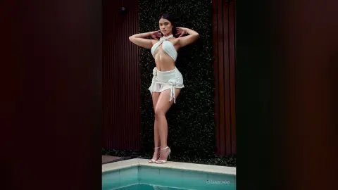 Elegant woman in a white halter crop top and mini skirt posing against a lush green garden wall.