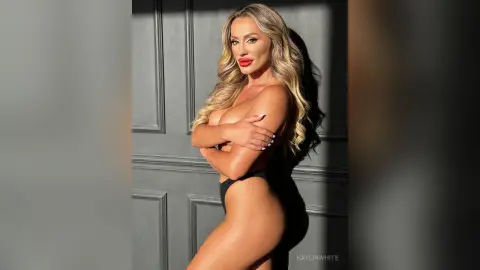 Glamorous blonde woman in black bikini posing against a dark paneled wall in warm sunlight.