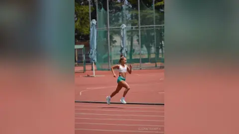 Athletic woman in white sports bra and green shorts running on an outdoor track under bright daylight.