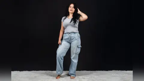 KaiaSinclair poses in a grey t-shirt and light blue cargo jeans against a black background.