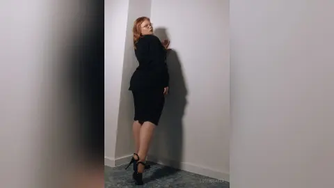 Redheaded woman in a chic black dress and heels posing against a white wall in a minimalist studio setting.