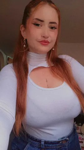 Redheaded woman in a white cutout long-sleeve top and denim jeans posing indoors with a soft mood.