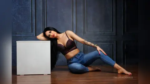 Glamorous woman in a black lace bra and blue jeans posing against a dark blue paneled wall.