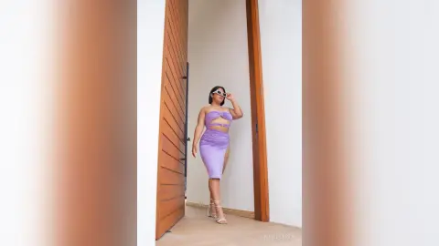 KimRoberth stands in a purple cut-out dress and clear heels against a white wall and wooden door frame.