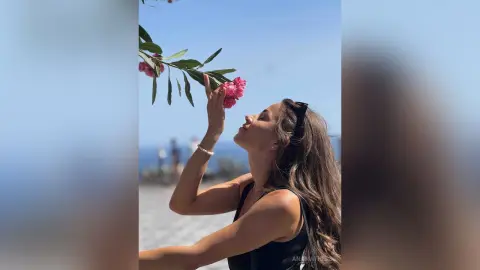 AnaMathers looks up while holding a pink flower near her face against a clear blue sky.