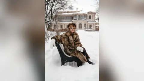 Elegant woman in a luxury fur coat and white gloves sitting on a sled in a snowy winter landscape.