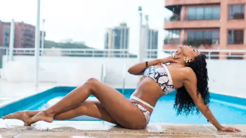 Curvy woman in a snakeskin bikini relaxing by a rooftop swimming pool on a bright summer day.